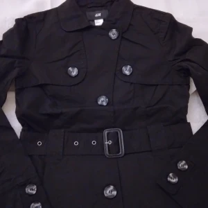 HELT NY & OANVÄND Svart fin dam jacka i trenchcoat stil. Strlk 34-36 / XS-S. - Helt NY & oanvänd fin svart jacka i trenchcoat-stil. Storlek: 34-36/XS-SMALL. Stängs med knappar och har avtagbart skärp i midjan, och har 2 fickor fram. Lapparna/tags sitter kvar sedan inköpet. Helt felfri/inga defekter.