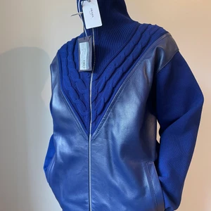 PRADA Kabelstickad Kofta, Leather Panel - PRADA 2019 blå kabelstickad samt läderpanel framtill, halv blixtlåst. Aldrig använd, taggar på.    Material: Polyester & Lammskinn    Storlek: EU 40, M   Mått: Axel mot skuldra - 47 cm   Midja - 53,5 cm  Längd - 67,5 (exkl. krage) Ärmlängd - 57 