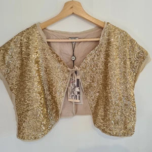 Noa Noa bolero jacka guldpaljetter Ny - Krämfärgad bolero med guldpaljetter. Märket är Noa Noa. Strl xxl. Ny med tags. Bystmått: ca 55 cm ×2 (ihopknuten). Mått från ärmhåla och till längst ner på boleron: ca 12 cm.