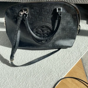 Guess Väska - Äkta Guess väska med axelband, i färgen grå/svart med gulddetaljier. Den är använd men det är inget man märker, känns som ny. Kommer med dustbag.
