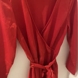 Omlottklänning - Vacker omlottklänning med knytband och dragkedja i sidan. En liten reva som syns på bild 3. Klänningen skimrar lite. Fin volang nertill. Lång ärm!