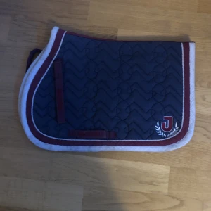 Schabrak  - Ett schabrak från Jacson i färg vit, röd marinblå. Lite smutsig. Säljs pga: kommer ej till användning. Storlek:ponny färg: marinblå, röd, vit. Säljs för 100 kr 