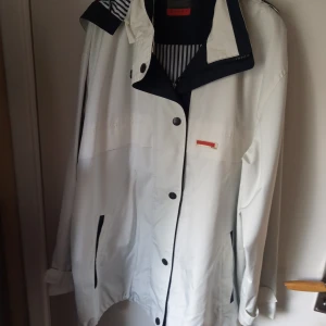  Vit sommarjacka med luva stl 44/46 - Fin sommarjacka med luva blåvit foder och knäppes med knappar och dragkedja .finns ett litet märke på ena ärmen annars i mycket gott skick,fickor fram.