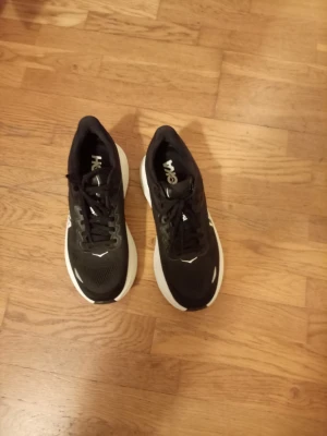 Svarta Hoka Bondi 9 sneakers  - Säljer ett par svarta Hoka Bondi sneakers 9 oanvända nya med vit tjock sula och svarta snören. Skorna har en sportig look med mesh-överdel och tydliga loggor på sidan och hälen. Perfekta för dig som gillar bekväma och snygga sneakers. 2399sek från butik kvitto finns