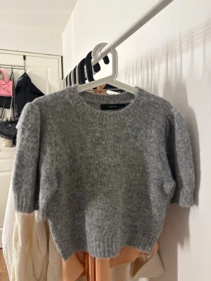 Grå stickad tröja från Vero Moda - Mysig grå stickad tröja från Vero Moda med korta ärmar och rund halsringning. Tröjan har en fluffig och mjuk struktur som ger en trendig look. 