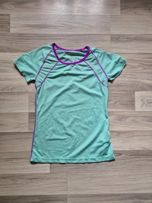 Mintgrön träningströja med lila detaljer - Snygg mintgrön träningströja med korta ärmar och lila kontrastsömmar samt lila kant vid halsen. Tillverkad i ett mjukt och stretchigt material som passar perfekt för träning eller sportaktiviteter. Figurnära passform och sportig look.