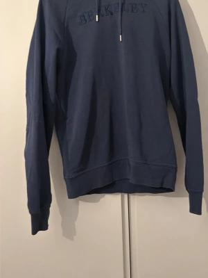 Blå hoodie med BERKELEY-tryck - Säljer en blå hoodie med huva och dragsnören. På bröstet står det 'BERKELEY' i ton-i-ton brodyr. Hoodien har långa ärmar, ribbade muddar och en klassisk passform. Perfekt för dig som gillar college-stil och vill ha något bekvämt och stilrent. ny pris 1700 mitt pris 600!