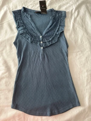 Blå ärmlös topp med volang - Supersöt blå ärmlös topp med volangdetaljer runt v-ringningen och små vita knappar framtill. Tyget har ett diskret mönster och toppen är lätt figursydd för en snygg passform. Perfekt till jeans eller kjol för en chill look.