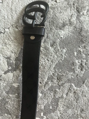 Svart bälte holografiskt tryck - Snyggt svart bälte med spänne i matt svart. Bältet har holografiskt mönster längs hela remmen och flera hål för justerbar passform. Perfekt accessoar för att lyfta din outfit. 116cm