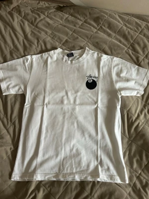 Vit Stüssy t-shirt med 8-ball print - Vit t-shirt från Stüssy med svart 8-ball print och logga på bröstet samt stort tryck på ryggen. Klassisk passform och rund hals. Tillverkad i mjuk bomull, perfekt för en avslappnad streetwear-look.
