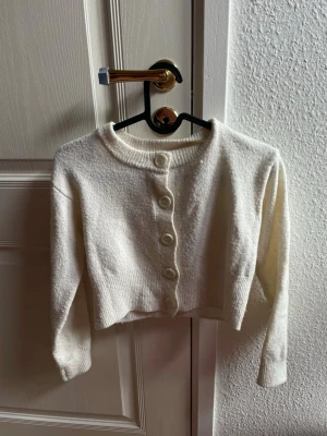 Vit cropped kofta från H&M 134/140 - Söt vit cropped kofta från H&M med stora knappar framtill. Koftan har rund halsringning, långa ärmar och är stickad i ett mjukt material. Perfekt att slänga över en topp för en chill look.