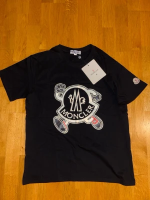 Svart Moncler t-shirt med tryck - Snygg svart t-shirt från Moncler med stort grafiskt tryck på bröstet och logga på ärmen. Klassisk rund hals och korta ärmar. Perfekt för dig som vill ha en trendig och bekväm look. Passar S och vissa M