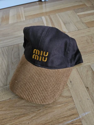Brun keps från Miu Miu med brodyr - Snygg brun keps från Miu Miu med gul broderad logga framtill. Skärmen är i ribbad manchester och själva kepsen är i slät bomull. Perfekt accessoar för en trendig och avslappnad look.