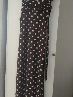 Brun lång prickig klänning - Lång brun klänning med vita prickar och volangdetalj vid halsringningen. Klänningen har ärmlös design och ett knytband i midjan för en snygg siluett, knytbandet går att justera✨
