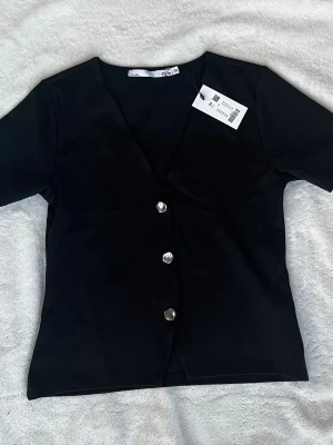 Svart v-ringad topp med knappar - Stilren svart topp med v-ringning och tre silvriga knappar framtill. Kortärmad modell i mjukt material, perfekt att matcha med jeans eller kjol. Helt ny och oanvänd med prislapp kvar, aldrig använd.
