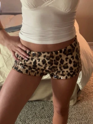 Leopardmönstrade paljettshorts - Säljer ett par korta shorts med leopardmönster och glittriga paljetter. Shortsen har låg midja och är i beige och svart med en tight passform. Perfekta för dig som vill sticka ut och gillar djurmönster och glitter.