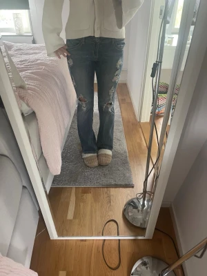 Blå slitna bootcut jeans med hål - Säljer ett par blå bootcut jeans med slitningar och hål på både fram- och baksida av benen. Jeansen har en klassisk femficksmodell och råa kanter vid hålen för en edgy look. Perfekta för dig som gillar en avslappnad och trendig stil.