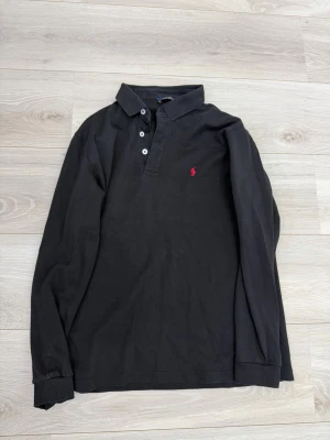 Svart långärmad polo Ralph lauren med röd logga - Svart långärmad polo Ralph lauren med klassisk krage och tre knappar framtill. På bröstet finns en broderad röd logga med polospelare. Skjortan är i mjuk bomull och har en clean, stilren look som passar till det mesta.