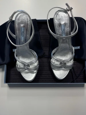 Silvriga pumps med knutdetalj från Prada - Snygga silvriga pumps från Prada med smala remmar och elegant knutdetalj framtill. Skorna har öppen tå, hög smal klack och justerbar ankelrem. Tillverkade i glansigt metallicläder som ger en lyxig känsla. 0689