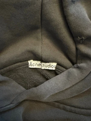 Svart hoodie Acne Studios Stockholm oversized - Säljer en svart hoodie från Acne Studios med trycket 'Acne Studios STOCKHOLM 1996' på ryggen. Hoodien har huva och är gjord i mjukt, fleecefodrat material för extra komfort. Perfekt för dig som gillar streetwear och vill ha en stilren look.