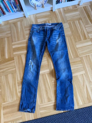 Blå slitna jeans med raka ben - Säljer ett par blå jeans med raka ben och slitningar på både fram- och baksida. Jeansen har klassisk femficksmodell och tydliga slitdetaljer på låren och knäna för en trendig, avslappnad look. Materialet är denim i bomull.