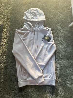 Ljusgrå hoodie från Stone Island - Säljer en ljusgrå hoodie från Stone Island med dragkedja framtill, två fickor och justerbar huva med snören. Klassisk Stone Island-patch på vänster ärm och logga på bröstet. Tillverkad i mjukt bomullsmaterial för skön känsla.