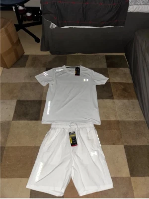 Vitt träningsset från Under Armour - Säljer ett fräscht vitt träningsset från Under Armour med t-shirt och shorts. Båda plaggen har logga på bröstet och benet, samt sportig passform. Perfekt för gymmet eller löprundan. Materialet är lätt och andas bra.