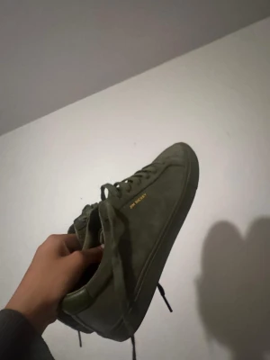 Olivgröna sneakers från Jim Rickey - Snygga olivgröna sneakers från Jim Rickey i mocka med matchande skosnören och diskret guldfärgad logga på sidan. Klassisk låg modell med rund tå och svart sula. Perfekta för dig som gillar stilrena och tidlösa skor.