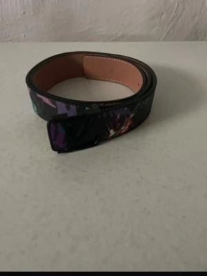 Svart bälte med färgglatt mönster - Snyggt svart bälte med färgglatt abstrakt mönster och svart spänne. Bältet har en stilren design och insidan är i beige. Perfekt accessoar för att lyfta din outfit.