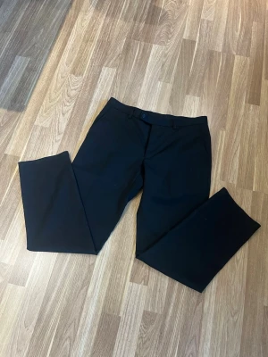 Svarta kostymbyxor med rak passform - Snygga svarta kostymbyxor med rak passform och bälteshällor. Tillverkade i en mjuk mix av 55% polyester och 45% ull som ger en klassisk look. Perfekta för dig som vill ha en stilren och enkel byxa till garderoben. Storlek 32-30