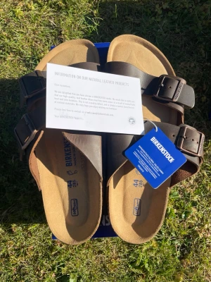 Oils lether  Birkenstock Arizona sandaler - Klassiska Birkenstock Arizona sandaler i brunt läder med två justerbara remmar och metallspännen. Fotbädden är ergonomiskt utformad och har en mjuk innersula. Yttersulan är i kork och gummi för skön komfort. Perfekta för en avslappnad och stilren look. De är riktigt populära och vid snabb affär så sänker jag priset, säljs billigt av en anledning 