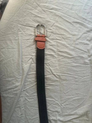 Svart flätat bälte - Snyggt svart flätat bälte med bruna skinn-detaljer och silverfärgat spänne. Perfekt accessoar för att lyfta din outfit och funkar till både jeans och chinos. 