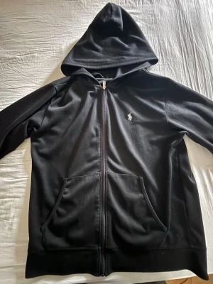 Svart hoodie från Polo Ralph Lauren - Snygg svart hoodie från Polo Ralph Lauren med dragkedja framtill och klassisk logga på bröstet. Hoodien har två stora fickor och en skön huva. Perfekt för en chill och stilren look. Materialet känns mjukt och bekvämt.