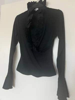 Svart Ruffle Blus - Sjukt snygg och cool blus.   Många fina detaljer. Omlott, v-ringad, detaljer längs ramen, fina band och tyg vid slutet på ärmarna.   Längd: ca 57 cm  Polyamid och elastisk i material. Se sista bilden.