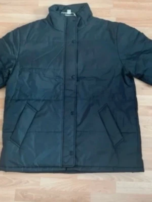 Ny. Black Puffer Jacket - This is a black puffer jacket with a high collar and snap button closure. It features two slanted front pockets and a quilted design for insulation. The material appears to be water-resistant, making it suitable for cold and possibly wet weather.