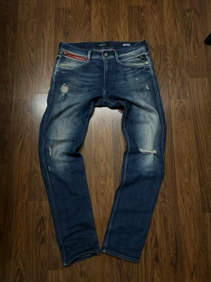 Replay jeans (Blå) - Säljer ett par blå jeans med slitningar och tvättade detaljer. Jeansen har smal passform och en röd dragkedjedetalj vid fickan. Perfekta för en avslappnad och trendig look. Skriv för storlek kunde inte välja men ni ser på bilden.