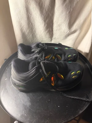 741 Jaylen Brown Basketball shoes Storlek 45 - Säljer ett par svarta sneakers med mesh-överdel och coola färgglada detaljer i orange, grönt och gult på sidorna och ovandelen. Skorna har snörning och en sportig, chunky sula som ger en modern look. Perfekta för dig som vill sticka ut med dina skor.