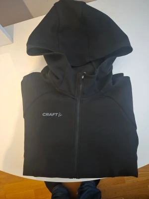 Svart hoodie med dragkedja från Craft - Snygg svart hoodie från Craft med hel dragkedja framtill och huva. Tröjan har diskret Craft-logga på bröstet och två fickor med dragkedja. Perfekt för chill eller träning. Materialet känns mjukt och sportigt.