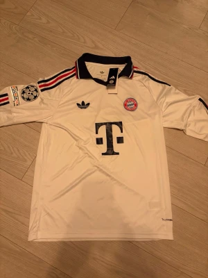 Vit Bayern München tröja långärmad - Vit långärmad Bayern München tröja från Adidas med svart krage och röda detaljer på axlarna. Klubblogga på bröstet, Champions League-märke på ärmen.