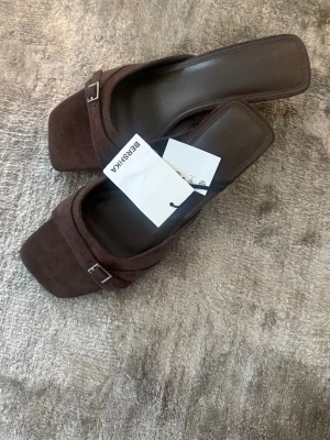 Bruna klackar med spänne från Bershka - Säljer ett par bruna klackar från Bershka med öppen tå och fyrkantig framdel. Sandalerna har en låg klack och dekorativa metallspännen framtill. Materialet ser ut att vara mockaimitation och de har en stilren, trendig look.