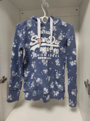 Blommig blå hoodie från Superdry - Snygg blå hoodie från Superdry med vita och ljusblå blommor över hela tröjan. Stor vit logga och text framtill, samt vita dragsnören i huvan. Mjuk bomullskänsla och känguruficka framtill. Perfekt för en chill och trendig look.