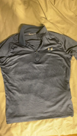  Under Armour golf tröjä - Snygg blå träningströja från Under Armour med halv dragkedja och ståkrage. Tillverkad i ett mjukt, stretchigt funktionsmaterial som andas bra. 
