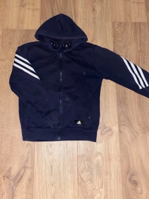 Mörkblå zip up från Adidas (S) - Snygg mörkblå hoodie från Adidas med dragkedja framtill och klassiska vita ränder på ärmarna. Tröjan har huva och ribbade muddar vid ärmslut och nederkant. Perfekt för en sportig och avslappnad stil.