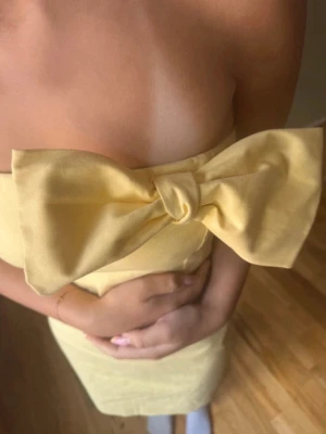 Gul axelbandslös klänning med rosett - Säljer en söt gul axelbandslös klänning med en stor rosett framtill. Klänningen är kort och har en slim passform som framhäver figuren. Perfekt för sommarens alla tillfällen!