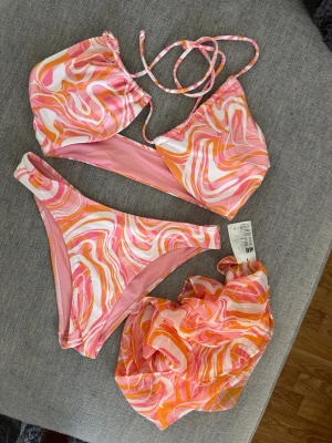 Färgglad swirl bikini från H&M - Säljer en bikini från H&M med swirlmönster i rosa, orange och vitt. Bikinin har trekantsöverdel med knyt och matchande trosa. Tillhörande sarong i samma mönster ingår. Perfekt för strandhäng och sommarvibes! Använd endast en gång. Överdel storlek 44. Underdel storlek 36
