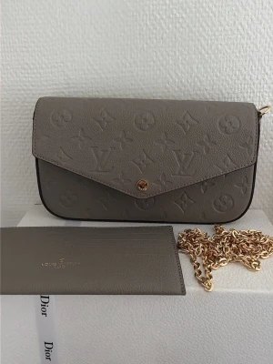 Beige Louis Vuitton kuvertväska - Säljer en beige kuvertväska från Louis Vuitton i präglat läder med det klassiska monogrammönstret. Väskan har en elegant, avlång form och stängs med en guldfärgad knapp framtill. Perfekt accessoar för dig som gillar lyxiga detaljer och stilren design.