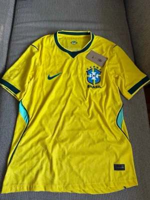 Brasilien matchtröja Nike gul - Säljer en officiell Brasilien matchtröja från Nike i starkt gul färg med gröna och blå detaljer. Tröjan har korta ärmar, rund halsringning med grön kant och CBF-emblemet på bröstet. Tillverkad i lätt och ventilerande funktionsmaterial.