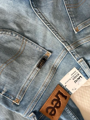 Ljusblå bootcut jeans från Lee - Säljer ett par ljusblå bootcut jeans från Lee med klassiska bakfickor och bruna sömmar. Jeansen har en avslappnad passform och är tillverkade i mjukt denimtyg. Perfekta för en casual och trendig look.