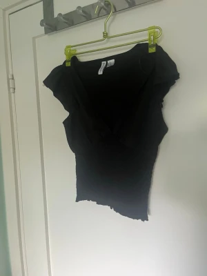 Svart v-ringad topp med volangärm - Snygg svart topp med v-ringning och korta volangärmar. Modellen är figurnära med smockad midja som ger en tight passform. Perfekt att styla med jeans eller kjol för en trendig look.