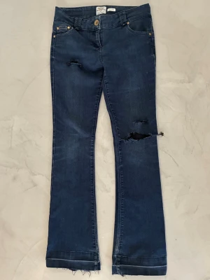 Mörkblå bootcut jeans med slitningar - Snygga mörkblå bootcut jeans med slitna detaljer och råa kanter nertill. Jeansen har klassisk femficksmodell, knappgylf och tydliga slitningar på låret och vid fickan. 